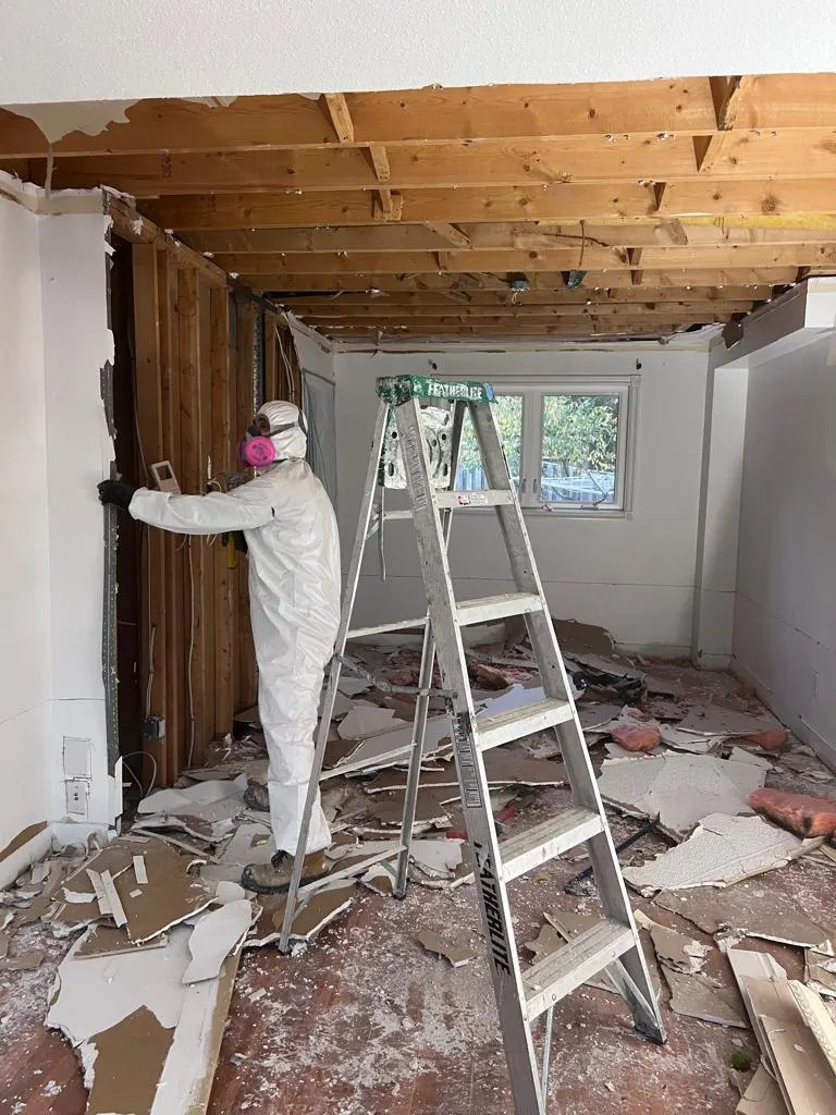 Technician in a full white coverall and respirator removing wall and ceiling drywall from a stepladder, with debris covering the floor below