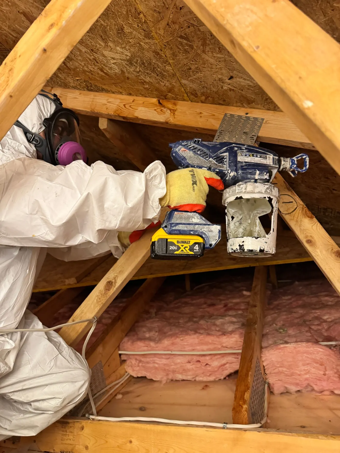 Technician in a white protective coverall and full-face respirator with pink P100 cartridges, working inside an attic with a spray applicator