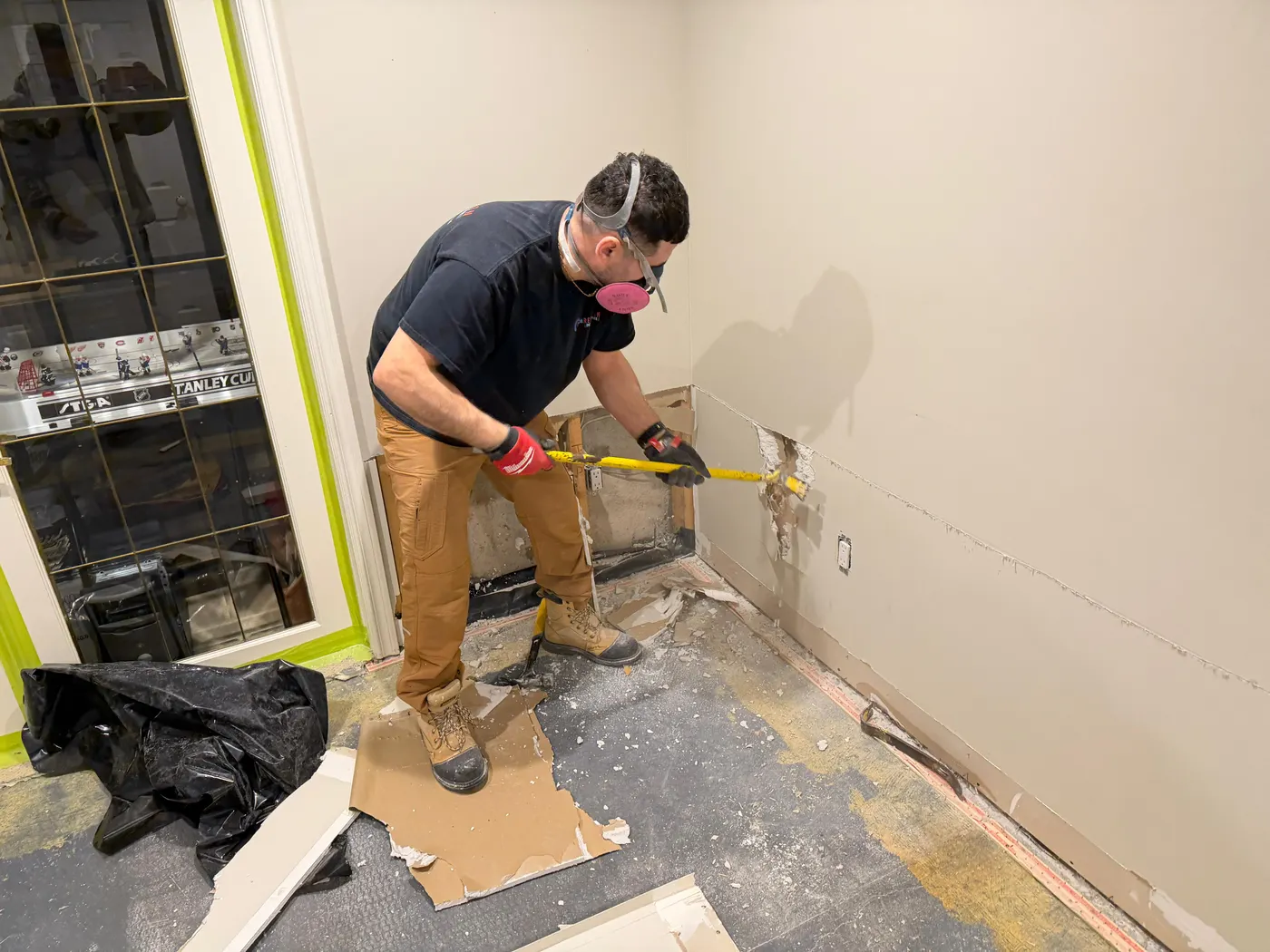 Technician in a respirator and work gloves using a pry bar to remove a section of water damaged drywall along a clean cut line