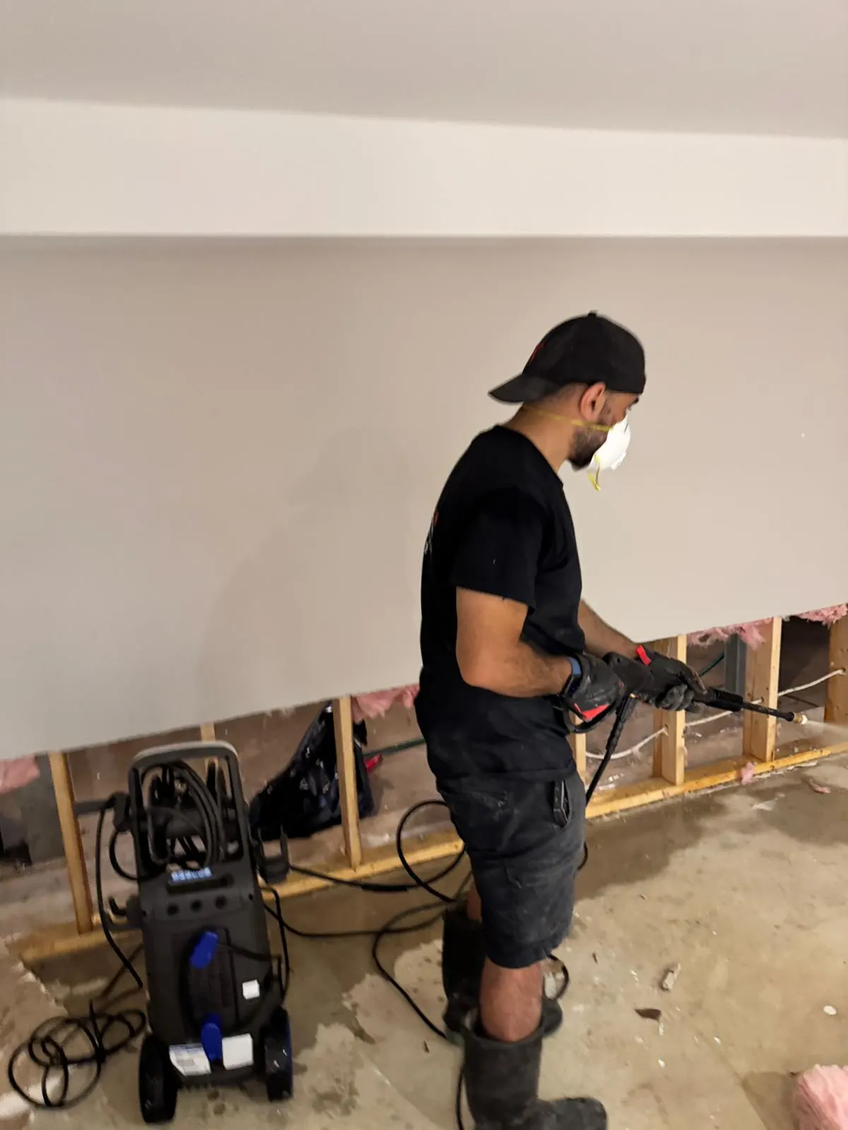 Technician in a mask and gloves working along an opened basement wall with a sprayer, insulation exposed between the studs