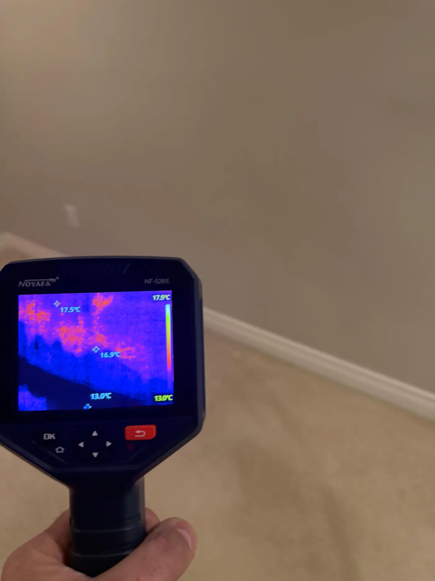 Thermal imaging camera held up to a wall, its screen showing a cold blue moisture pattern spreading behind the drywall above the baseboard