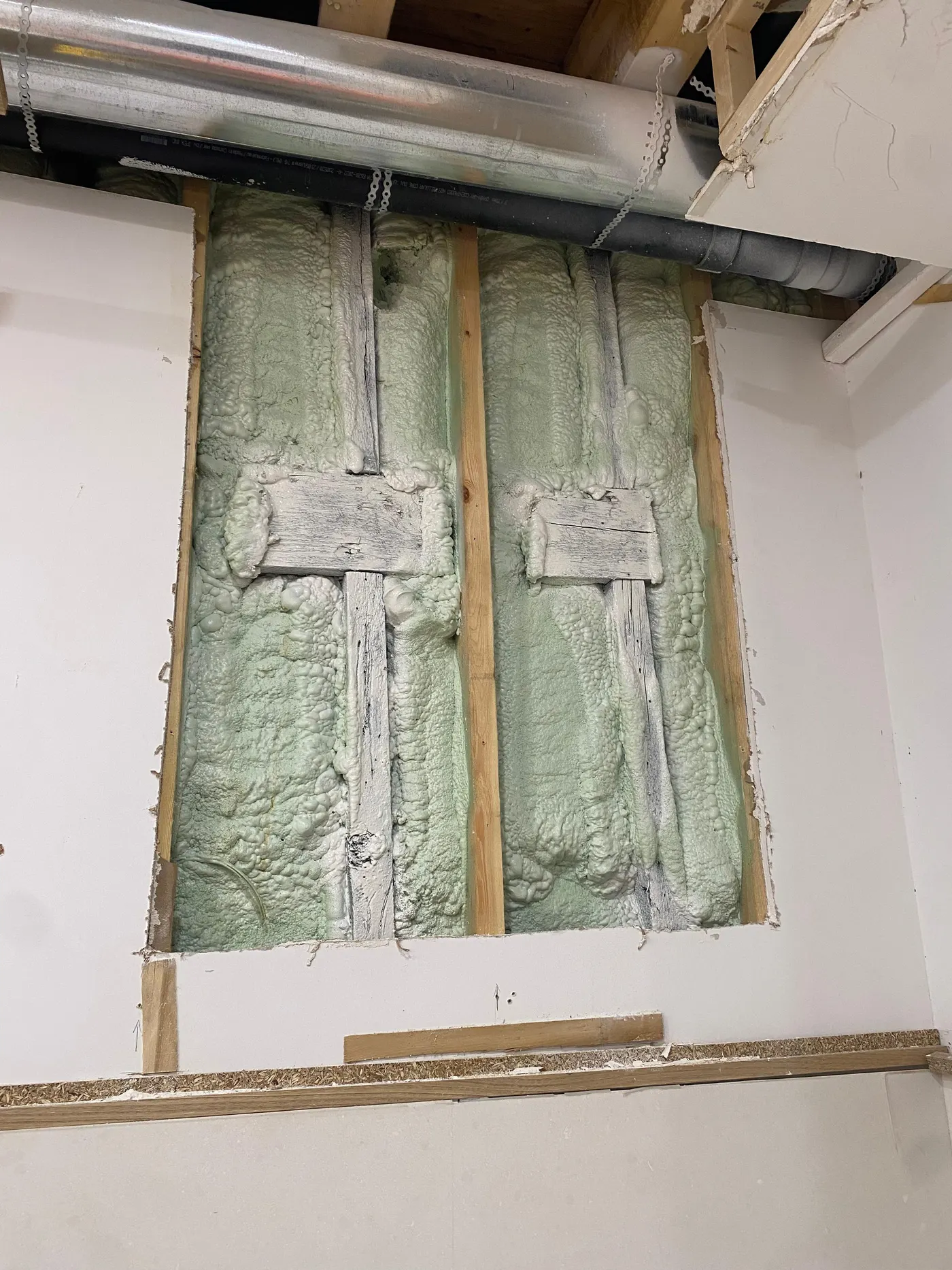 Wall cavity opened between studs to expose spray foam insulation and overhead ductwork, with the surrounding drywall cut back to sound material